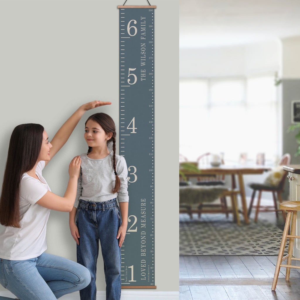 A personalised traditional style canvas height chart hanging on a wall, with a child standing on a chair to reach the highest measurement, and an adult assisting. The chart features measurement markings in centimeters and feet/inches.